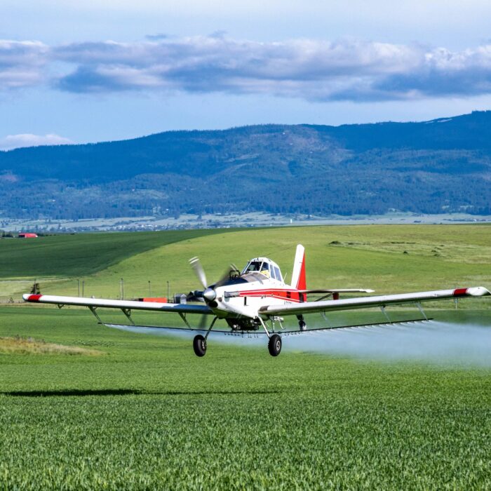 A small aircraft sprays pesticide over a lush green agricultural field with mountains and cloudy skies in the background. The plane flies low distributing the spray evenly across the crops