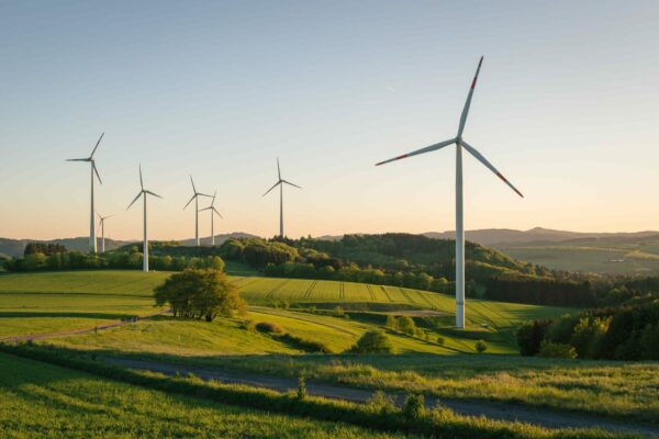 A scenic landscape with several wind turbines scattered across green rolling hills under a clear sky at sunset. The turbines are standing tall, and the fields are lush and vibrant, with a path winding through the grassy terrain. Hills and mountains are visible in the distance