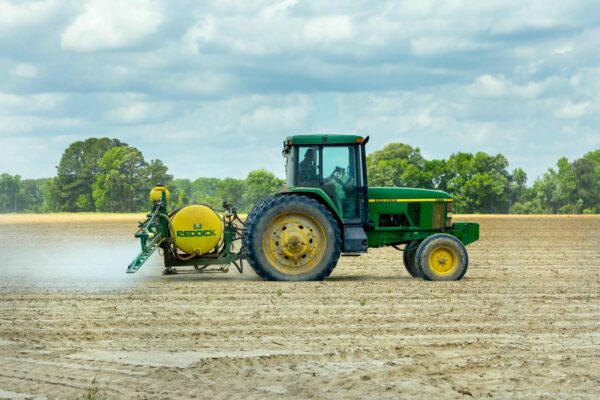 A green tractor pulls a yellow Redick sprayer attachment across a large partially tilled field under a cloudy sky The field is bordered by trees in the background