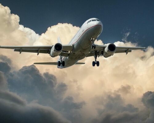 A commercial airplane with its landing gear lowered flies against a backdrop of dramatic, billowing clouds in the sky. The aircraft is captured from below showcasing its undercarriage and wings. The scene exudes a sense of motion and impending arrival.