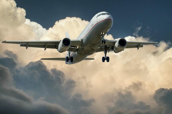 A commercial airplane with its landing gear lowered flies against a backdrop of dramatic, billowing clouds in the sky. The aircraft is captured from below showcasing its undercarriage and wings. The scene exudes a sense of motion and impending arrival.