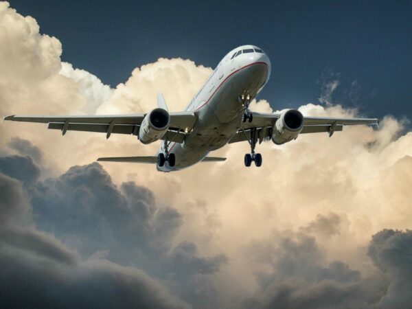 A commercial airplane with its landing gear lowered flies against a backdrop of dramatic, billowing clouds in the sky. The aircraft is captured from below showcasing its undercarriage and wings. The scene exudes a sense of motion and impending arrival.