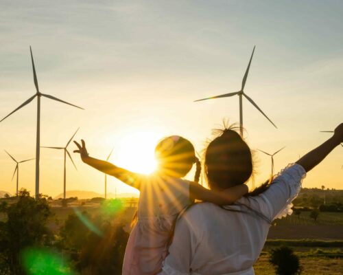 A person and a child standing in a field with wind turbines, embracing with the sunset casting a warm glow, arms raised toward the sky celebrating the scenic moment and green energy around them