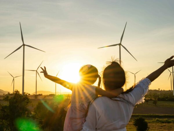 A person and a child standing in a field with wind turbines, embracing with the sunset casting a warm glow, arms raised toward the sky celebrating the scenic moment and green energy around them