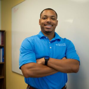 A man in a bright blue polo shirt stands confidently with arms crossed in front of a whiteboard Shelves with books are partially visible in the background He is smiling at the camera