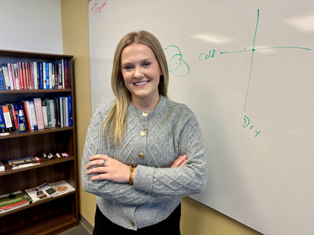 A person with long hair, wearing a gray sweater, stands with arms crossed in front of a whiteboard displaying a graph labeled cold and dry. A bookshelf filled with various books is behind them.