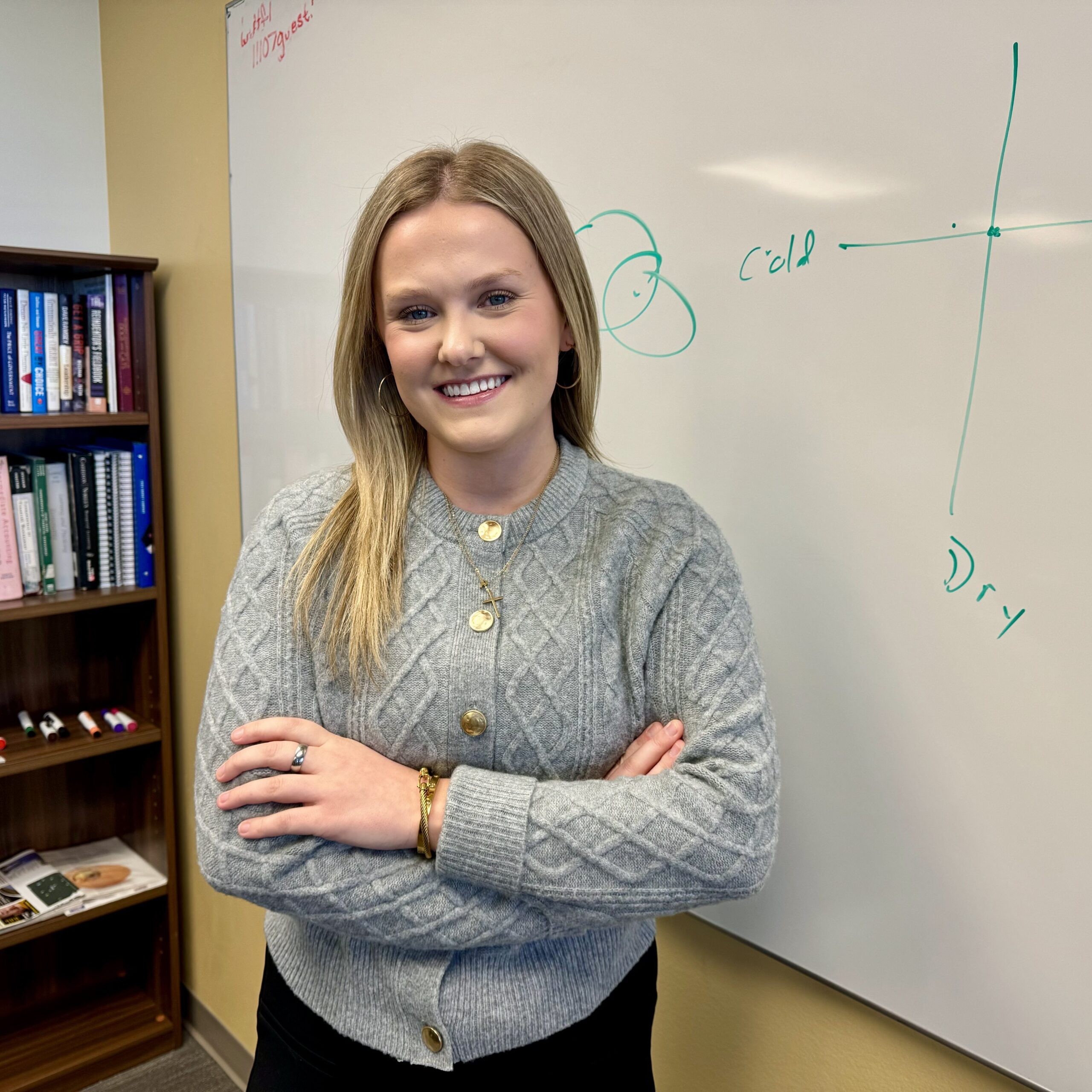 A person with long hair and a gray sweater stands smiling with arms crossed in front of a whiteboard displaying a diagram. A bookshelf filled with books is visible in the background.